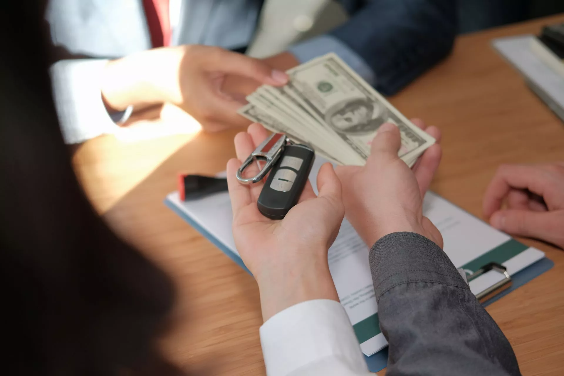 桌面上可見一人將一疊現金與汽車鑰匙交付給對方，畫面中雙手同時接觸金錢與鑰匙，背景有文件與資料夾，呈現交易、交付款項或完成購買手續的情境。