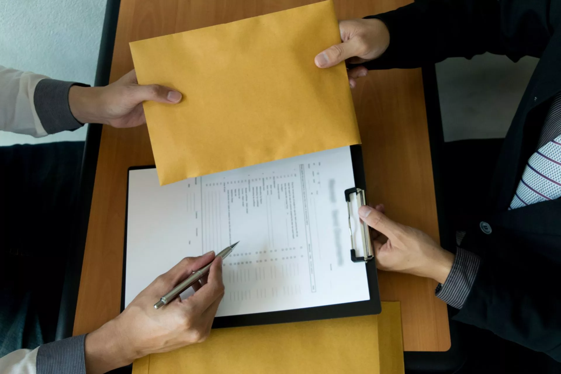 兩人於木質桌面上交接牛皮紙文件袋，下方夾板夾著合約文件，其中一人手持筆準備簽署的商務場景。