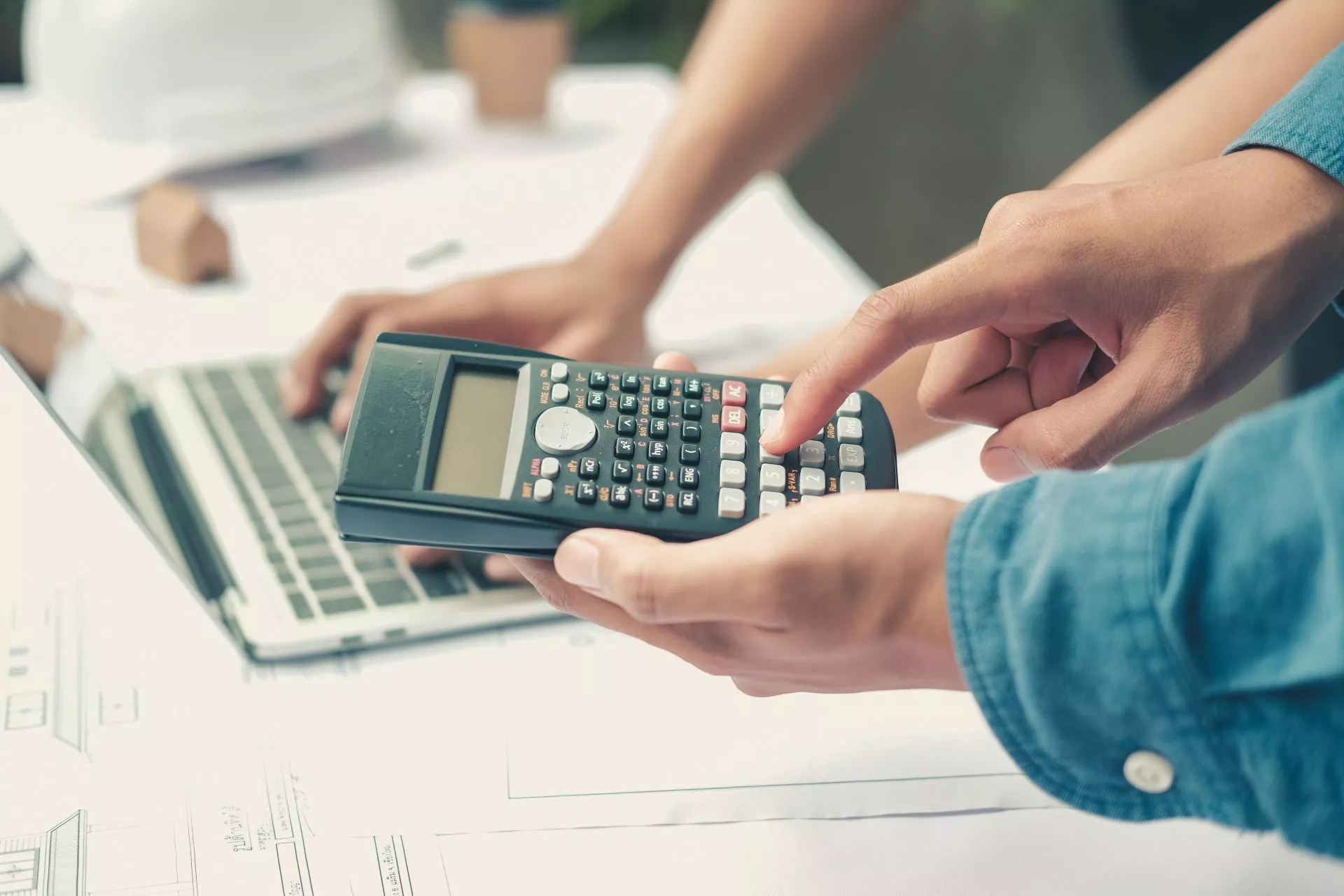 畫面中可見一人手持計算機按鍵進行計算，前方桌面攤開著文件與圖表，旁邊另有人操作筆電，呈現多人協作進行數據試算、預算評估或工程／財務規劃的工作情境。