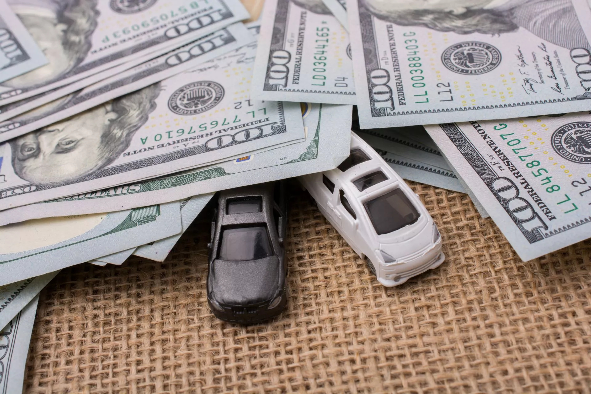 多張美元鈔票散放在桌面上，底下露出黑色與白色的小型汽車模型，象徵購車資金、車貸或汽車消費支出概念。