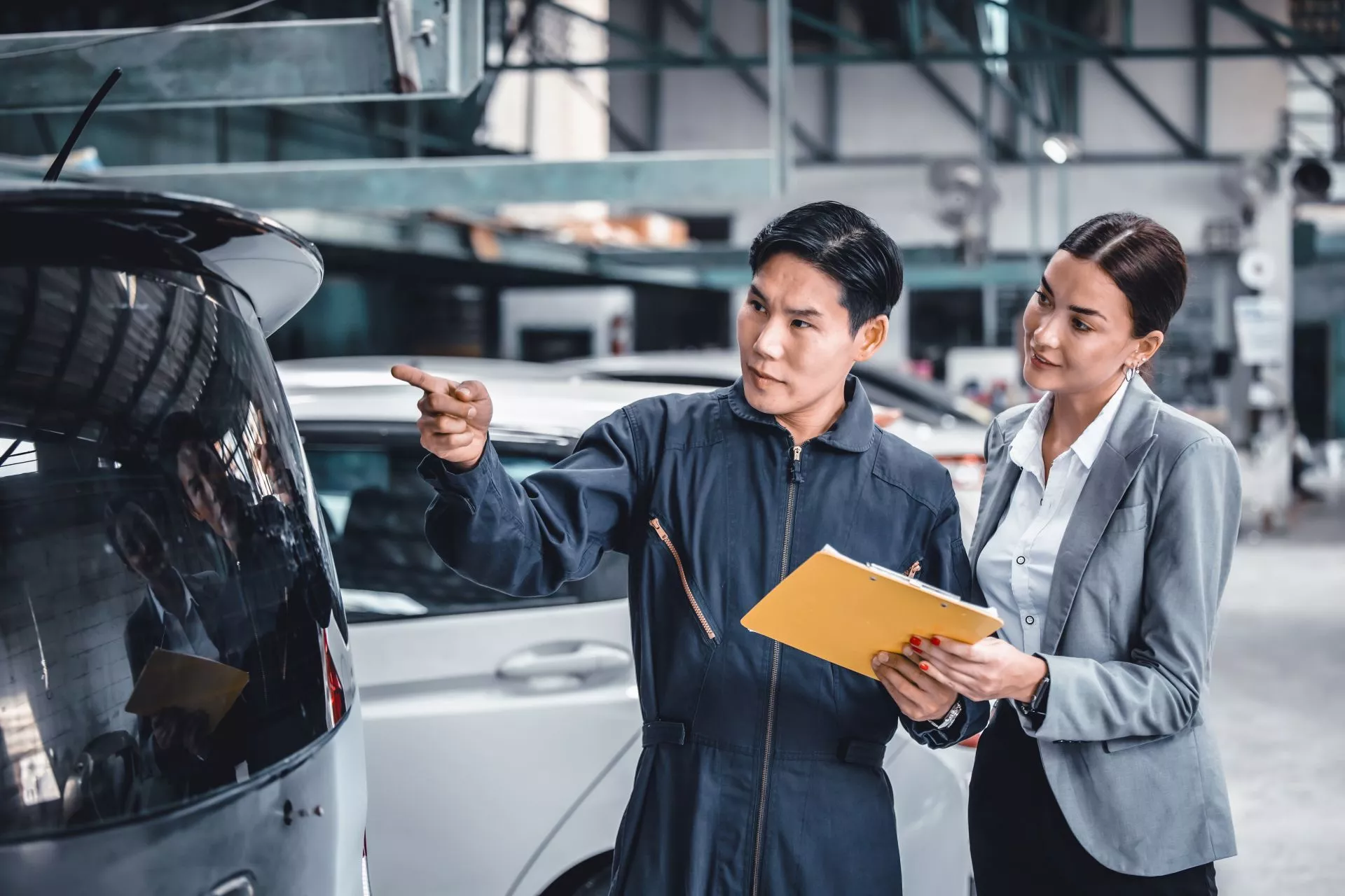 汽車維修廠內，技師指向車輛後方部位說明狀況，旁邊業務人員手持文件夾陪同確認車況。